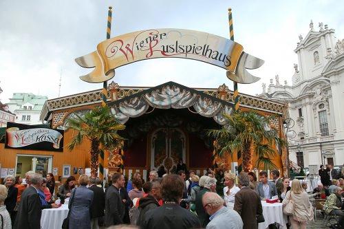 Foto vom Eingangsbereichs des Wiener Lustspielhauses: Eingangsportal mit zwei Palmen darüber ein gemalter Baldarchin, zwei Fahnenstangen und ein Schild mit dem Namen,  Besucher:innen stehen um Stehtische