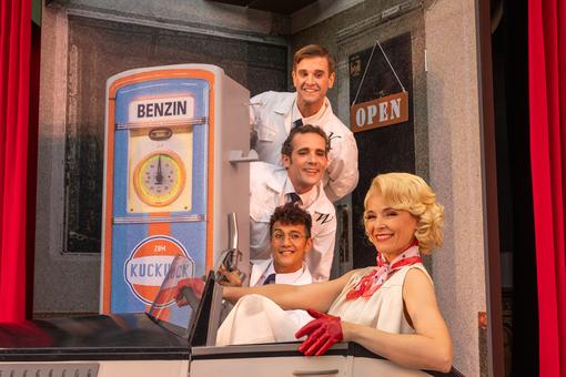 The scene shows three gentlemen / gas station attendants at the pump of a gas station in front of an elegant blonde lady in a convertible