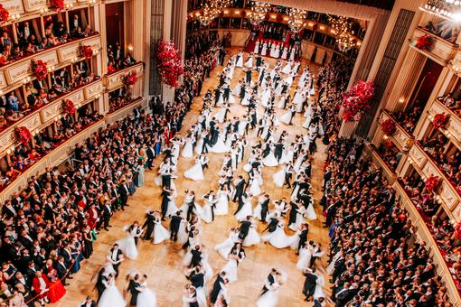 Zu sehen ist der festlich geschmückte Innenraum der Wiener Staatsoper, in den Logen und am Rande des Parketts Ballgäste, in der Mitte die Tanzpaare / Debütant:innen des Jungdamen- und Jungherrenkomitees