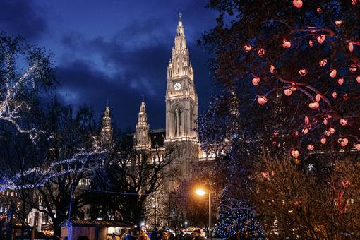 Das Foto zeigt das Wiener Rathaus in abendlicher Beleuchtung, im Vordergrund die festlich geschmückten Bäume des Rathausparks und einige Hütten des Wiener Christkindlmarktes