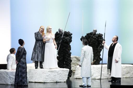 The scene photo from the opera “Das Rheingold” from the Ring of the Nibelungs shows the singers in black and white costumes on a light-colored stage. In the center is a blonde woman in a white dress, next to her a man with long red hair in a black coat
