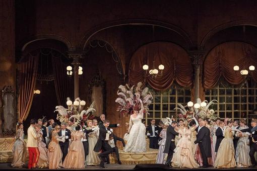 Szenefoto aus der Operette "Die Fledermaus", zeigt eine Abendgesellschaft in eleganten Kleidern, Smokings und Uniformen in einem festlichen Ambiente, in der Mitte die Hauptdarstellerin in einem weißen Abendkleid auf einem Podest stehend