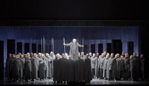 The scene photo from the opera “Götterdämmerung” from the Ring of the Nibelungs shows a crowd of people in gray uniform-like costumes, in the middle the main actor standing elevated above this crowd, all in front of a dark background
