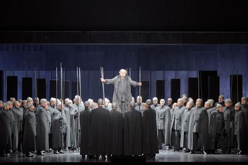 Das Szenefoto aus der Oper "Götterdämmerung" aus dem Ring der Nibelungen zeigt eine Menschenmenge in grauen uniformartigen Kostümen, in der Mitte der Hauptdarsteller erhöht über dieser Menge stehend, alles vor dunklem Hintergrund