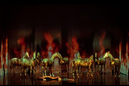 Scene from the opera “Walküre” at the Vienna State Opera. In the photo, a person is lying on the stage floor, a person with a spear in their hand is standing next to them, behind them golden horses against a black and red background