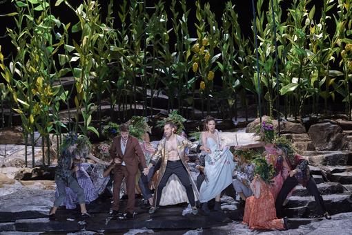 Das Szenefoto aus der Oper "Don Giovanni" zeigt den Hauptdarsteller, der eine Frau und einen Mann an der Hand hält, umgeben von 6-8 Personen mit Pflanzenschmuck auf dem Kopf, im Hintergrund ein Wäldchen aus Pflanzen