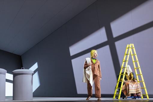 Das Szenefoto aus der Oper "Fin de Partie" zeigt grauen Bühnenraum, rechts steht eine gelbe Holz-Stehleiter, darunter sitzt ein verschleierte Frau im Rollstuhl, links davon ein Mann in braunem Anzug und grellgrünem Hemd und mit gelbem Haar
