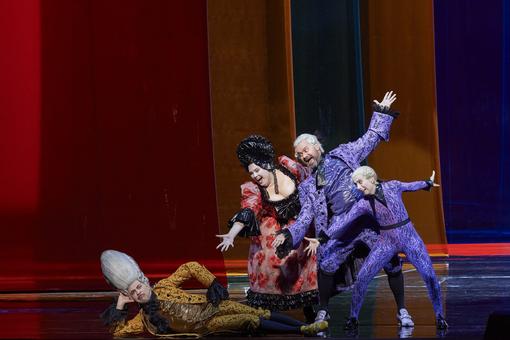 The scene photo from the opera “Il barbiere di Siviglia” shows four actors, a man in a yellow historical costume with a high wig is lying on the floor, two actors dressed in bright purple and a woman in a sweeping dress are pointing at him