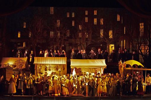 The scene photo from the opera “La Boheme” shows an evening scene at a market in yellow light, a large crowd of people in historical costumes crowding in front of the stalls, trees and tall houses with illuminated windows in the background