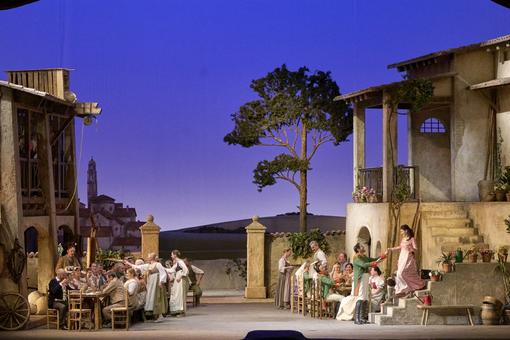 The scene shows a party in an inner courtyard of a country estate in the south, numerous guests are seated around two tables, the lady of the manor strides down the outside steps of the house and is greeted by a soldier