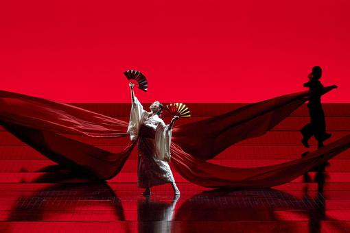 Szenefoto der Oper Madama Butterfly, die Hauptdarstellerin in traditionelle japanischer Kleidung mit zwei Fächern in Händen im Scheinwerferlicht vor tiefrotem Hintergrund