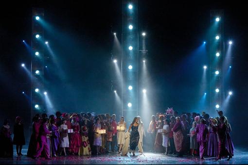 The scene photo from the opera “Roméo et Juliette” shows a colorfully dressed party, in the middle the leading actress in a dark dress and long brown hair