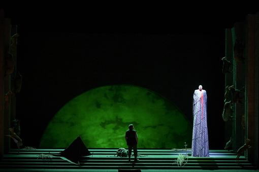 The scene photo from the opera “Siegfried” from the Ring of the Nibelungs shows a stage illuminated in black and green, a person standing in the dark and looking at an oversized, brightly lit figure in a floor-length purple striped costume
