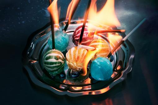 The photo shows colorful lollipops lying on a silver tray and burning in front of a dark background