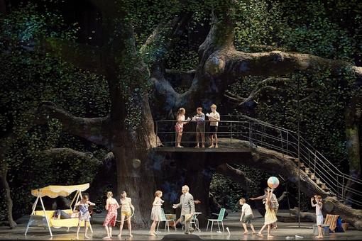 The scene photo from the opera “Werther” shows an old spreading tree with three children on a thick branch, with more children playing in front of it, camping furniture and a yellow Hollywood swing