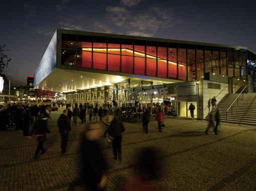 Foto von der beleuchteten Halle F der Stadthalle am Abend, davor Menschen, die in die Halle strömen