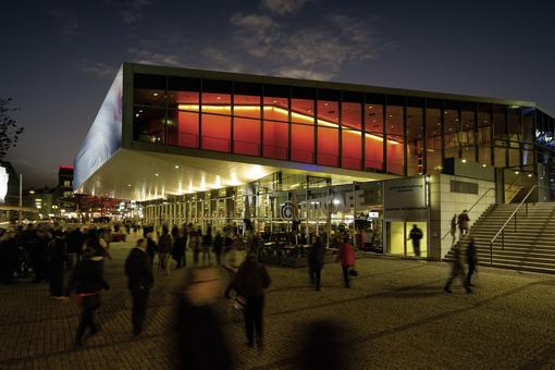 Photo of the illuminated Hall F of the Stadthalle in the evening, in front of it people streaming into the hall