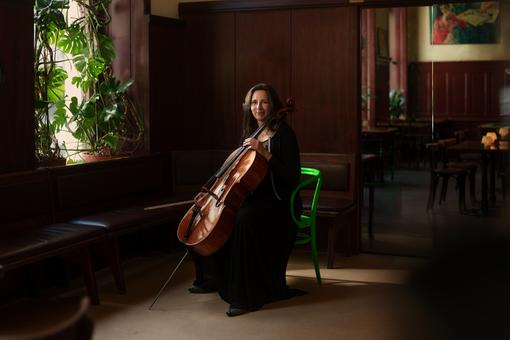 Photo of symphonist Maria Grün, sitting on a green armchair in a Viennese pub and holding her cello in her hands