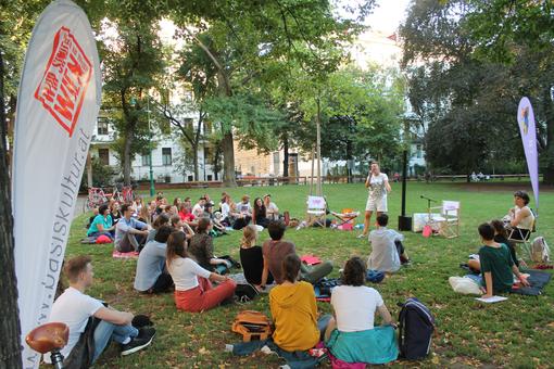 Scene in a Viennese park, artist with microphone speaks or sings, audience sitting on the grass in front of it