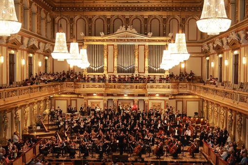 Zu sehen ist der goldene Saal des Musikvereins, ein Jugendorchester auf der Bühne musizierend, Publikum seitlich und auf den Rängen 