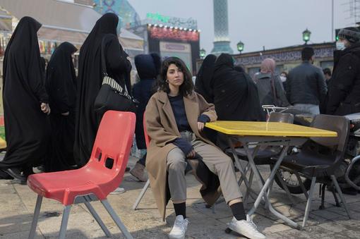 Photo of young woman dressed in western causal and sitting at a table, behind her Muslim women with black hishab and floor length black coats walk by