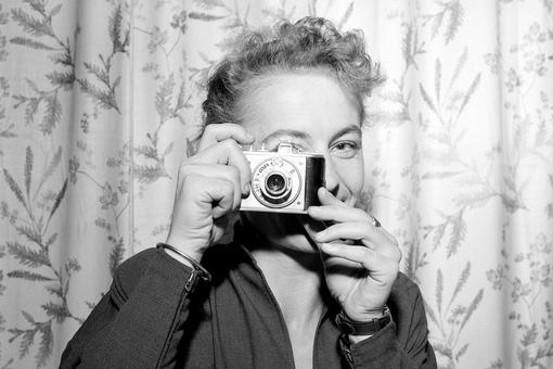 The black and white photo shows a woman holding a historical camera in front of her right eye and turning towards the viewer as if she were taking a photo