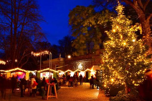 Foto eines Adventmarktes mit beleuchteten Hütten und einem beleuchteten Christbaum im Vordergrund, in abendlicher Stimmung
