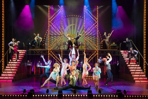 Scene photo of the show with dancers in costumes from the 1920s, colorful lighting, illuminated show stairs on both sides