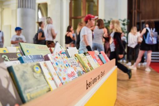 The photo shows a shelf with various children's books in the foreground and children of different ages in the background
