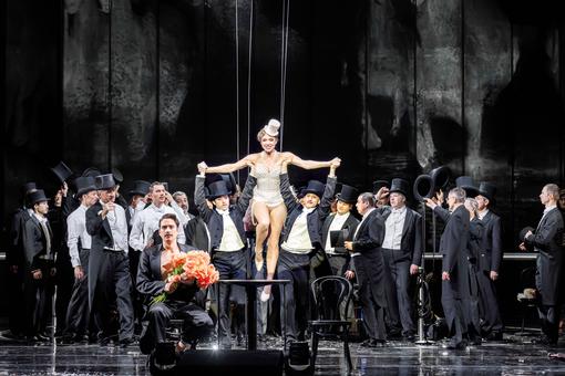 The photo shows the leading actress in a white pearl jersey with a white hat surrounded by a group of men in tails and top hats, in front of them a man with a red-orange bouquet of flowers