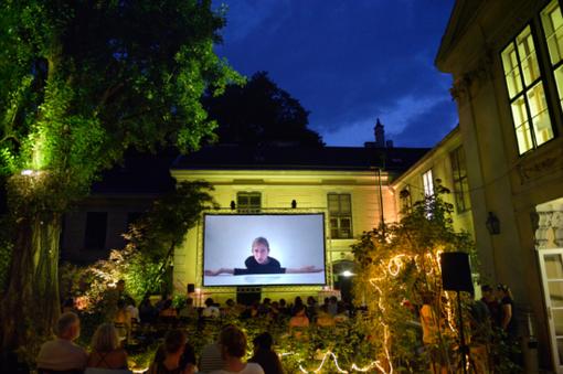 Evening scene: big screen in a courtyard, audience in front, trees on the side