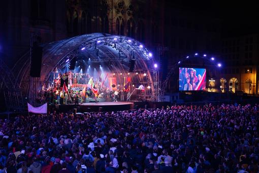 The photo shows the illuminated stage at the opening of the Wiener Festwochen with the artists and musicians on stage and the large audience in front of them