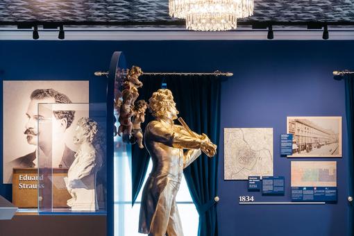 Exhibition view with a golden statue of Johann Strauss, a bust of the composer, a large-format photo, etc. in a room with deep blue walls