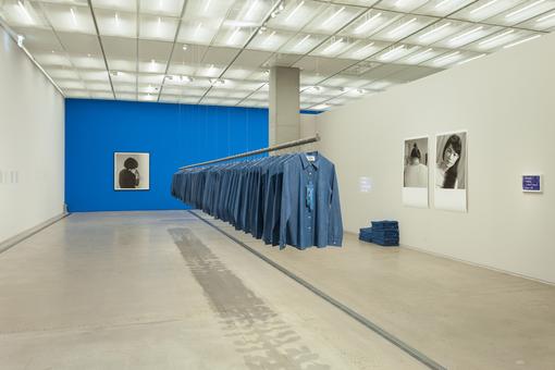 Exhibition view: countless medium blue suits hanging on a free-floating rod in a room with two white and one blue wall