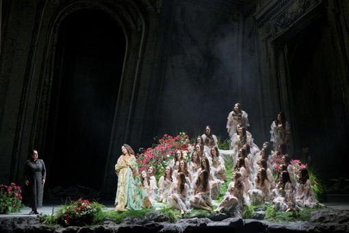 The photo shows the leading actress in a long coat with a floral design in front of a group of many women who look like fairies sitting in a meadow, with a stern woman in all black on the right looking at the group of women
