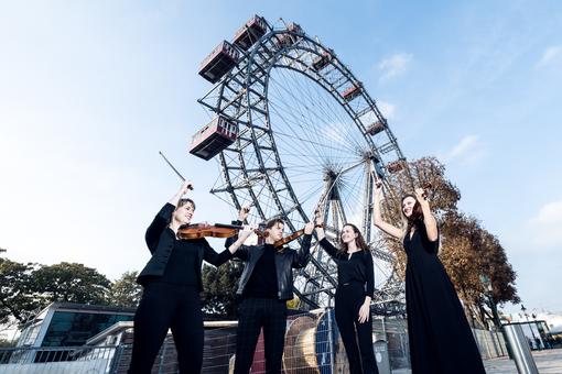 The photo shows four violin players dressed in black in front of the Vienna Giant Ferris Wheel