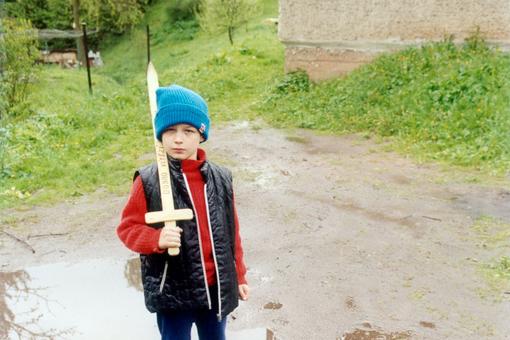 Das Foto zeigt einen Buben in rotem Pullover, darüber ein schwarzes Gilet, mit blauer Wollmütze und einem Holzschwert in der Hand, das er an seine Schulter lehnt. Er steht im Grünen, vor einer Pfütze auf nassem braunem Boden 