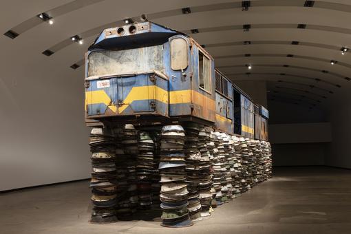 Die Ausstellungsansicht zeigt eine blaugelbe, alte und völlig heruntergekommene Lokomotive, die auf unzählige bunten, rostigen Metallschüsseln aufgebockt ist und wie auf dem Schüsselberg gestrandet wirkt