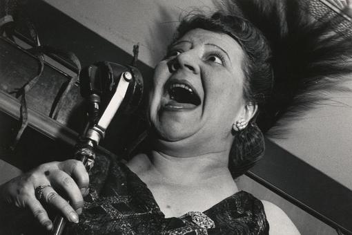 The black-and-white photograph by Lisette Model shows a singer at Cafe Metropole, New York City, in 1946. She is holding a microphone in her hand, her mouth wide open.