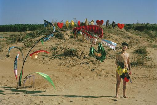 Foto einer bunten Installation, die eine Art Palme darsellen soll und eines jungen Mannes in Badehose inmitten eiiner kleinen Sanddüne