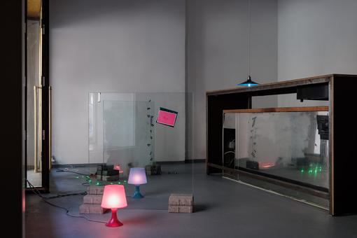 Photo of an installation view: in a high room with gray walls, on the floor of which are different colored small lamps and strings of lights, bricks holding upright panes of glass.