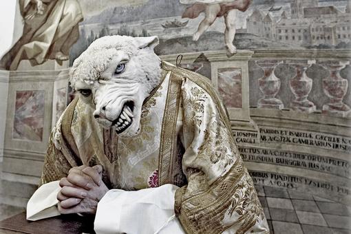 Das Bild zeigt ein Schaf mit Wolfschnauze und Wolfgebiss, im Gewand eines hohen katholischen Würdenträgers, an einem Gebetsstuhl gekniet und die Hände gefaltet 