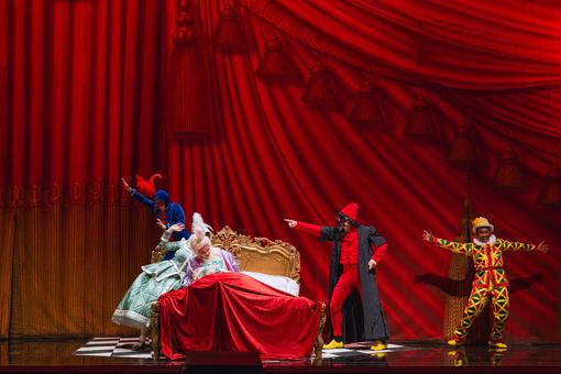Das Szenefoto aus der Oper "Le nozze di Figaro zeigt vier Hauptdarsteller:innen in bunten Kostüme, die sich um ein barockes Bett mit roter Überdecke tummeln, vor tiefrotem Hintergrund, der wie ein Vorhang mit Falten und Quasten aussieht