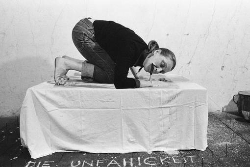 Black and white photo of Vallie Export kneeling bent over on a platform with white cloth with her mouth biting off a piece of an elongated object