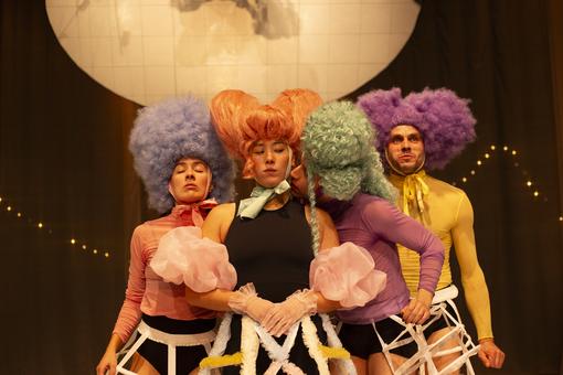 The photo shows the four main actors with big colorful wigs and colorful costumes, huddled close together, eyes closed, a man trying to kiss a woman