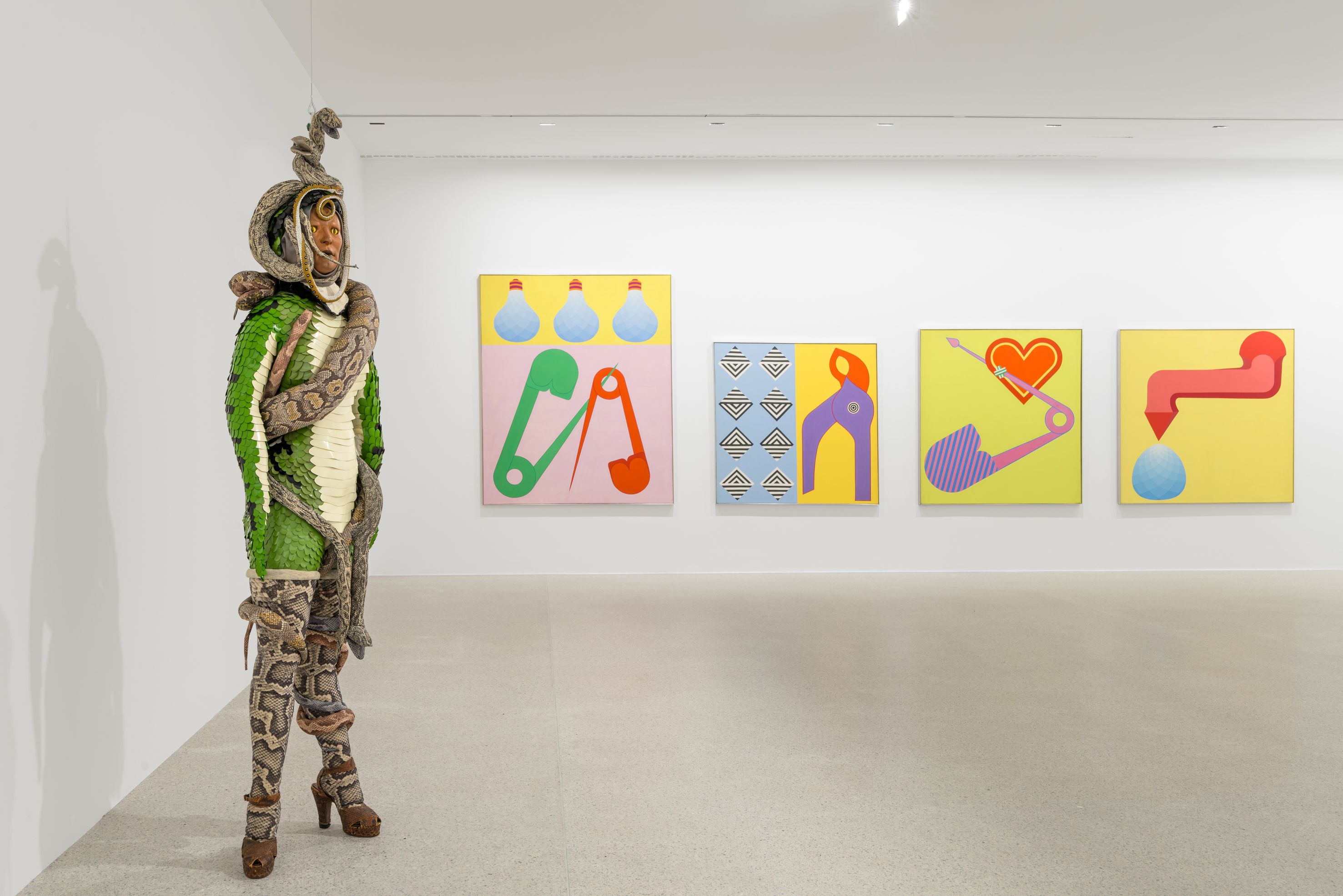 Ausstellungsansicht: Mapping the 60s. Kunst-Geschichten aus den Sammlungen des mumok / Sine Hansen @ Foto: Klaus Pichler / mumok Exhibition view: in the foreground a figure in snakeskin and entwined with snakes, in the background four colorful, bright paintings with everyday objects, e.g. pliers, light bulbs