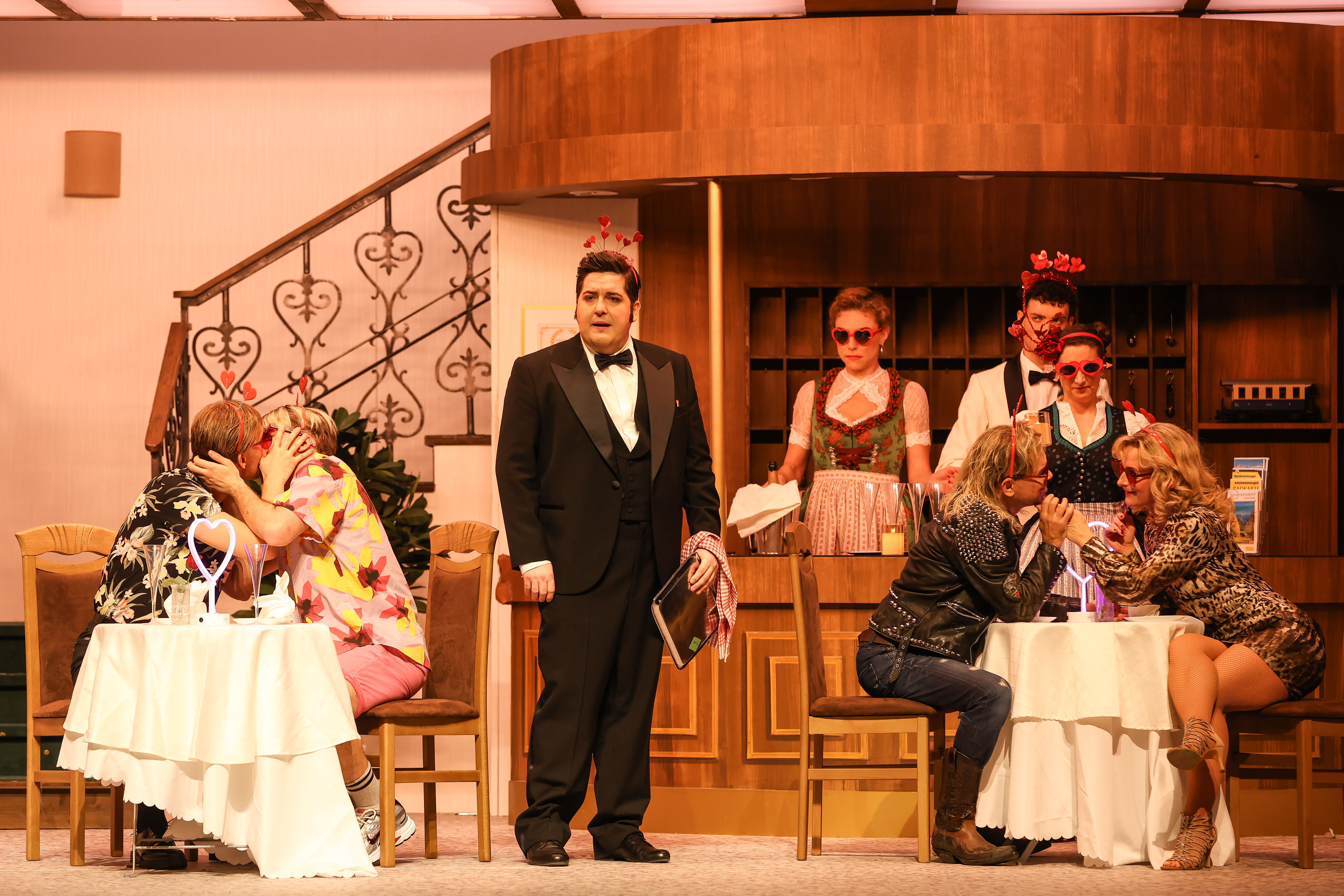 Volksoper Wien / Im weißen Rössl: Jakob Semotan (Leopold Brandmeyer, Zahlkellner), Annette Dasch (Josepha Vogelhuber, Wirtin), Christoph Stocker (Der Piccolo), Ensemble © Foto: Barbara Pálffy / Volksoper Wien Scene photo from the play “Im weißen Rössl” in front of a hotel reception in the 1950s, where a woman in a dirndl and a waiter are standing, in front of two tables with flirting couples, in between the head waiter speaking to the audience