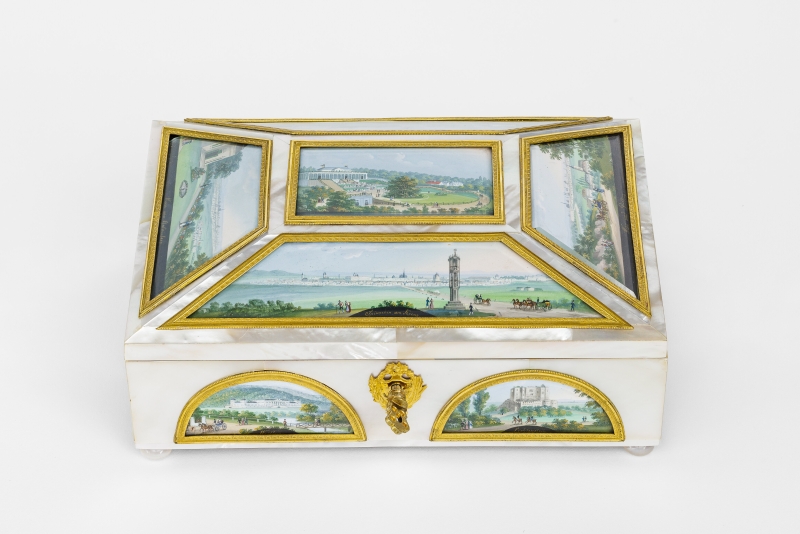 Ausstellung: Wien, Wien nur du allein. Wigand – Alt – Oláh © Heidi Horten Collection, 2025 Das Foto zeigt eine weiße Schatulle mit goldenem Schloss und Schlüssel und aufwendigen Email-Einlegearbeiten mit goldenen Rahmen, die unterschiedliche Wiener Motive zeigen. Der erhöhte Deckel zeigt fünf Einlegearbeiten mit Perlmutt unterlegt.