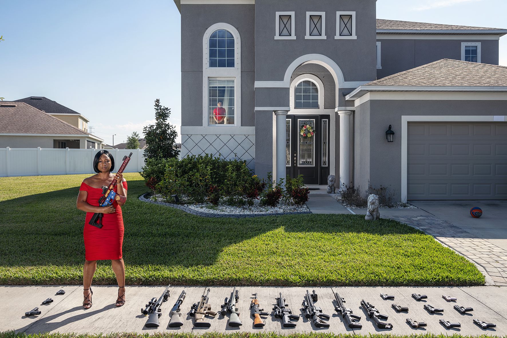 Gabriele Galimberti: Avery Skipalis (33) – Tampa, Florida, 2021, aus der Serie The Ameriguns © Foto: Gabriele Galimberti The photo shows a large gray one-story house in a suburb in Florida. In front of it stands a dark-skinned woman in a red dress with a rifle in her hands. Next to her in the front yard of the house are another 10 rifles and 10 pistols.