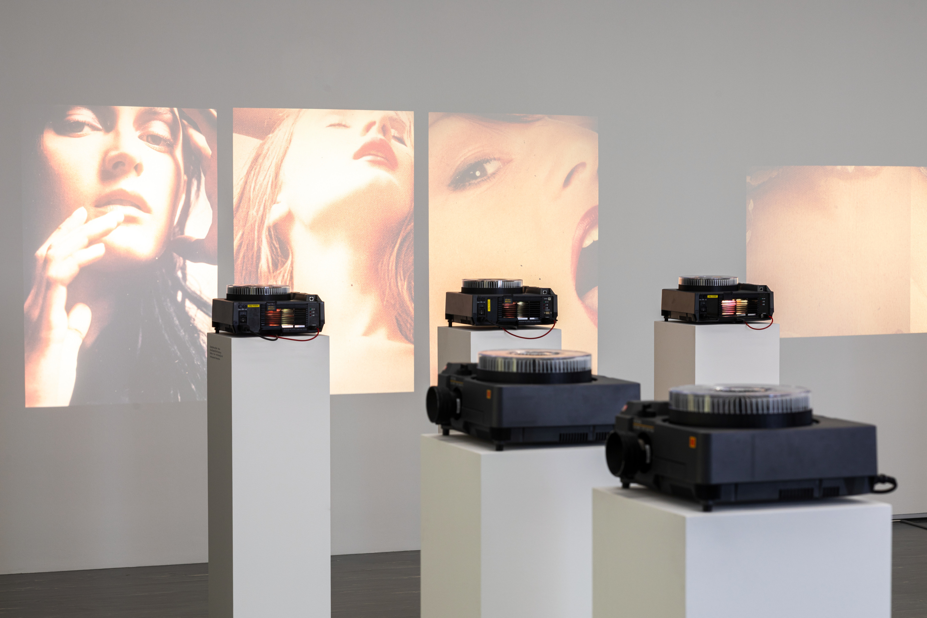 Ausstellungsanischt Maria Hahnenkamp, Belvedere 21© Foto: Johannes Stoll / Belvedere, Wien The photo shows an exhibition view of “Maria Hahnenkamp” at Belvedere 21, with five slide projectors on white steles that continuously project photos of women onto the wall